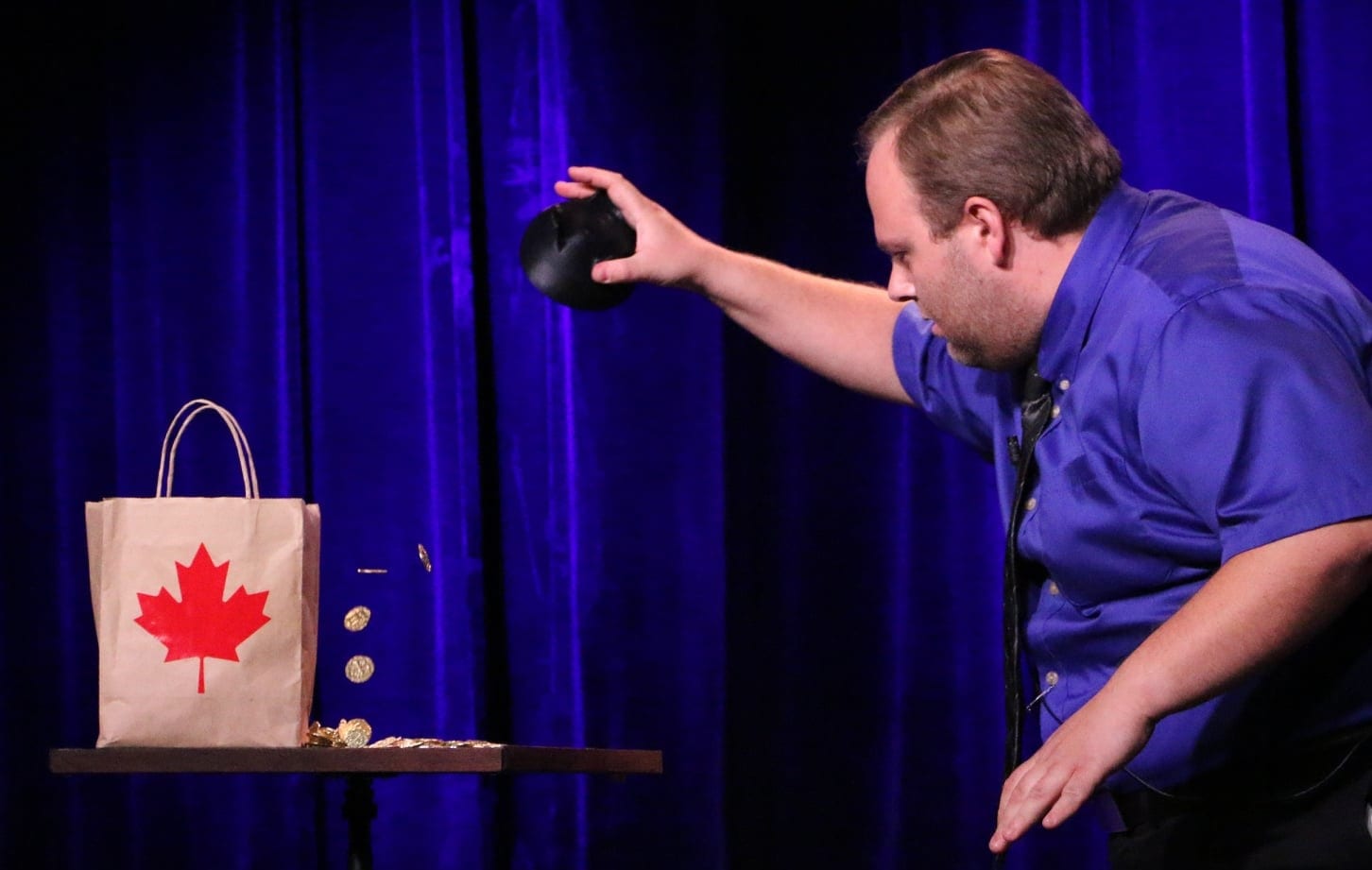 Hanneman the Magician pouring gold coins from a hat toward a maple-leaf bag on a blue-lit stage